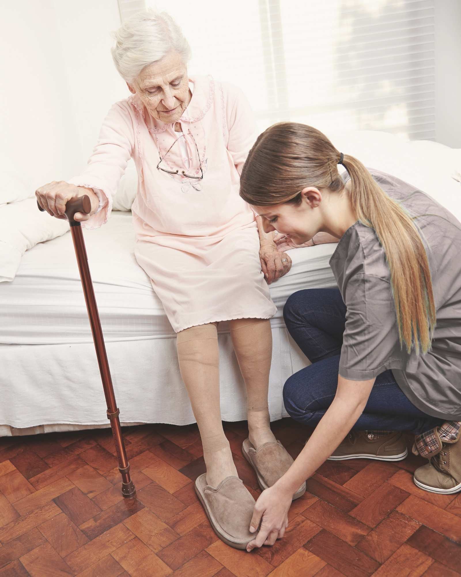 A caregiver helps an older adult put on slippers beside a bed, offering gentle support in a home setting.