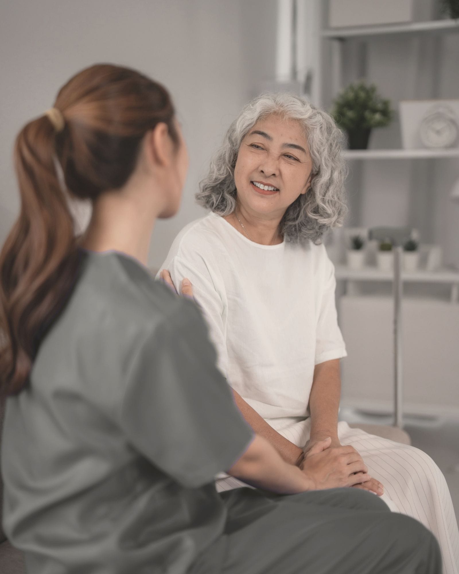 A caregiver in scrubs chats warmly with an older adult in a bright, welcoming home environment.